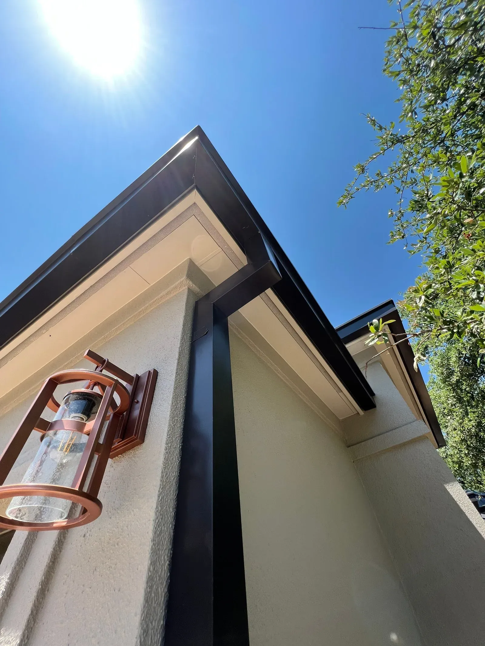 Dark seamless metal gutter and downspout along a stucco roofline against a clear sky