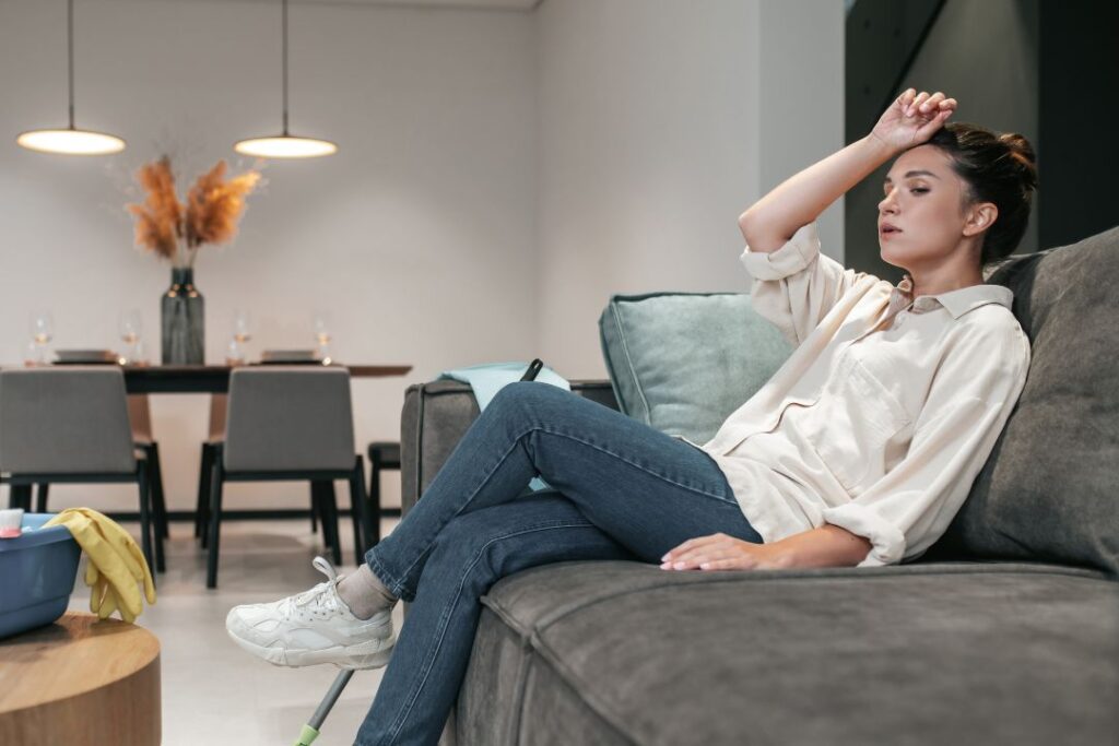 Tired woman resting on a couch with cleaning supplies nearby in a modern living room.