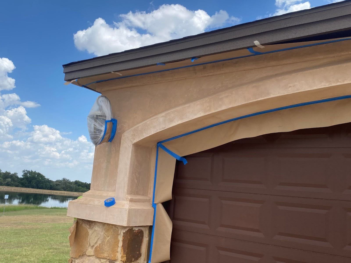 A close-up of a house's exterior being painted. Blue painter's tape is used to mask off the trim and garage door.