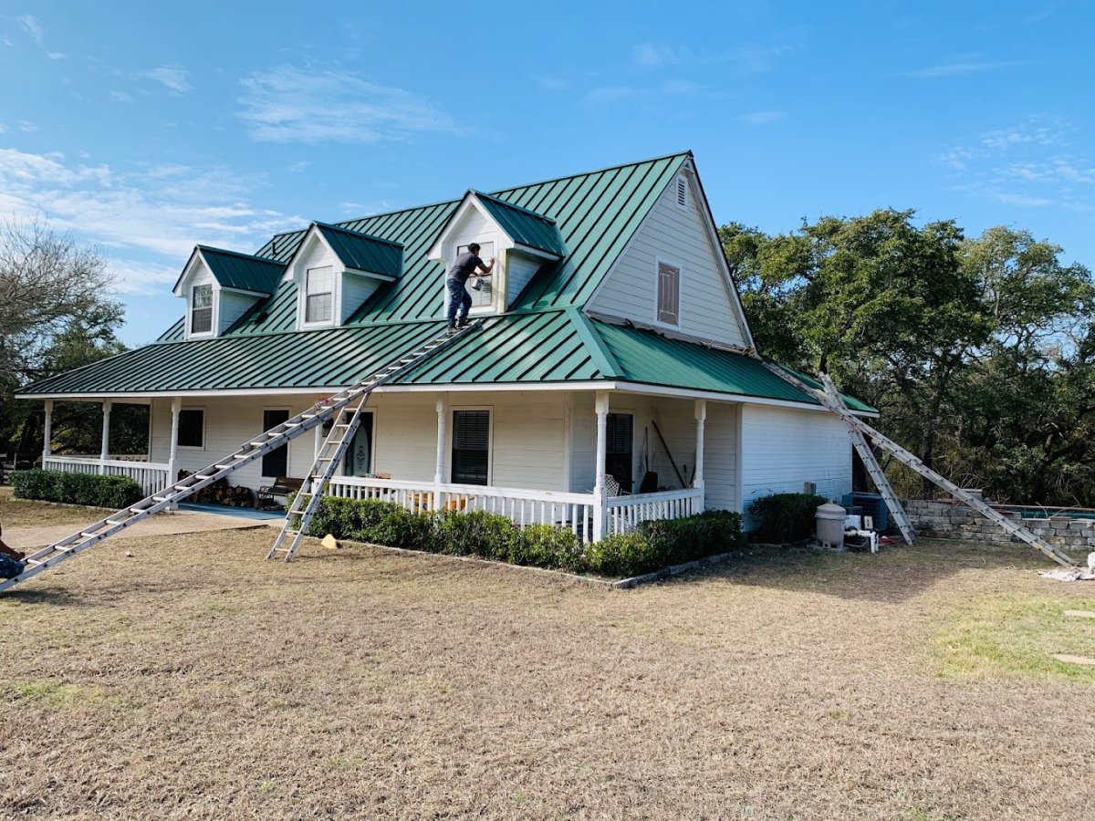 A large house with a green metal roof and white siding. A worker is on the roof, painting. Ladders are leaning against the house.