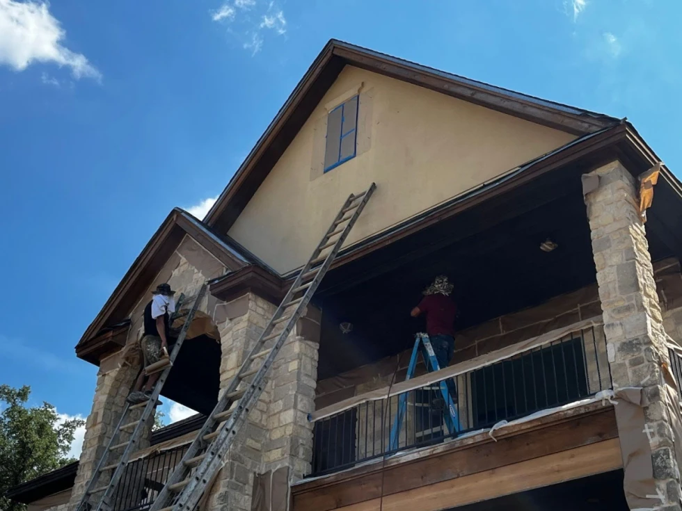 Two workers on ladders are painting the exterior of a house with beige stucco walls and a stone facade.