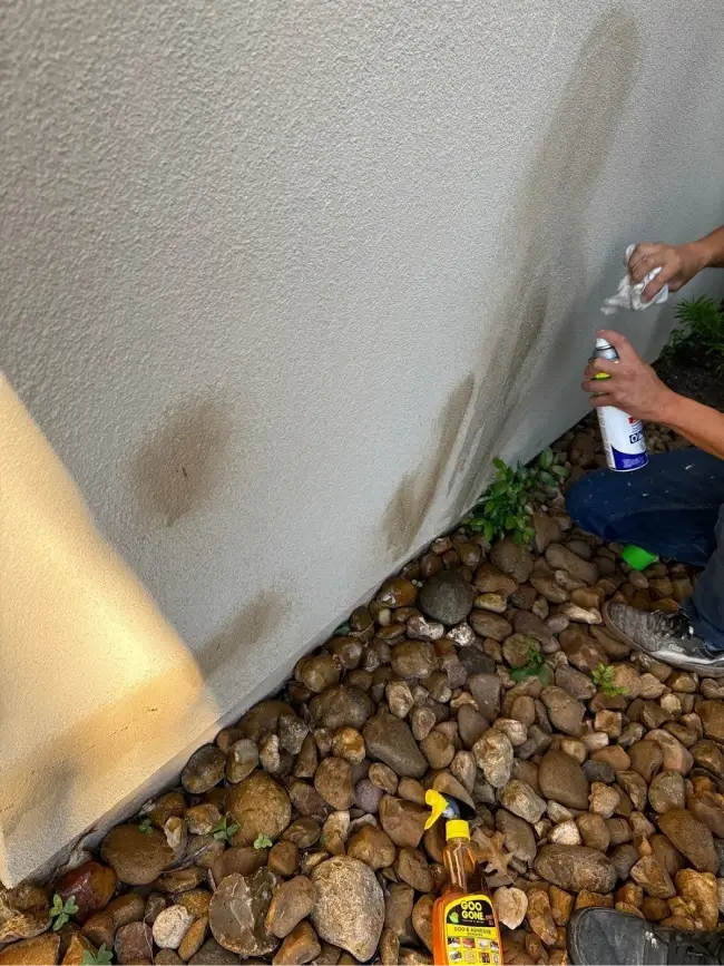 A close-up of a person cleaning stains from a light-colored wall using spray cleaners and a cloth.