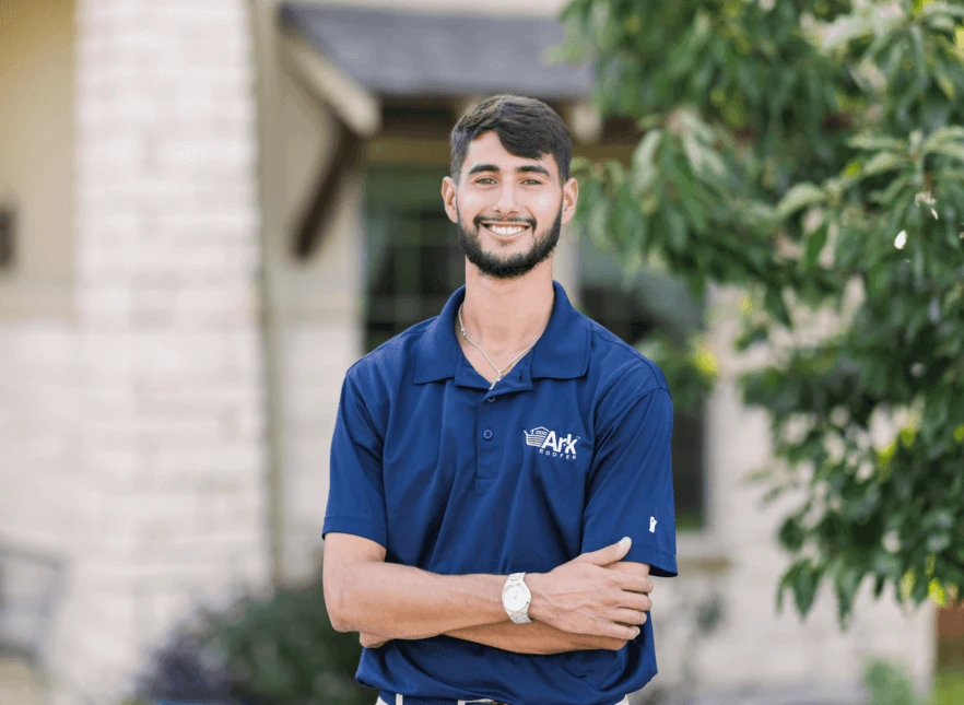 A man smiling, standing outside, wearing a blue Ark Roofing polo shirt.