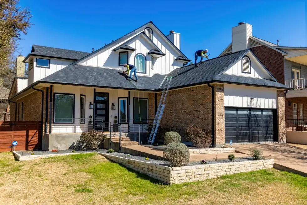 Workers installing roofing on a modern two-story home with white siding and brick accents